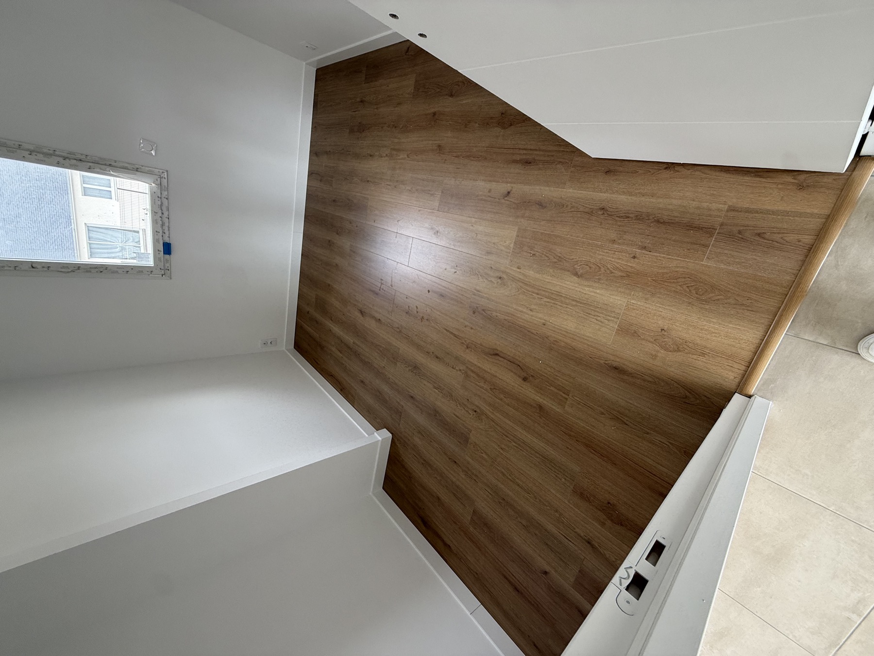 Parquet aspect bois posé dans une petite pièce blanche, vu depuis le seuil carrelé par Aristopeintre